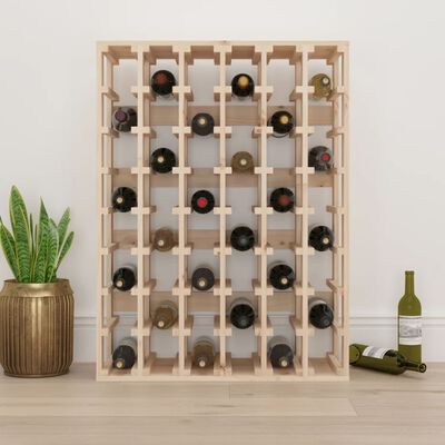 Wooden wine rack filled with wine bottles against a white wall with plant and wine bottles nearby.